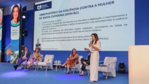 Foto da procuradora-geral Cibelly Farias palestrando no evento. Ela está em pé, falando ao microfone, no palco, que é uma estrutura azul. Atrás dela, os demais participantes, sentados em poltronas um ao lado do outro. Nas laterais, há dois telões que projetam a sua foto e nome. No telão do meio, é projetada a apresentação. À frente dos palestrantes, parte do público, de costas para a foto.