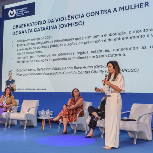 Foto da procuradora-geral Cibelly Farias palestrando no evento. Ela está em pé, falando ao microfone, no palco, que é uma estrutura azul. Atrás dela, os demais participantes, sentados em poltronas um ao lado do outro. Nas laterais, há dois telões que projetam a sua foto e nome. No telão do meio, é projetada a apresentação. À frente dos palestrantes, parte do público, de costas para a foto.